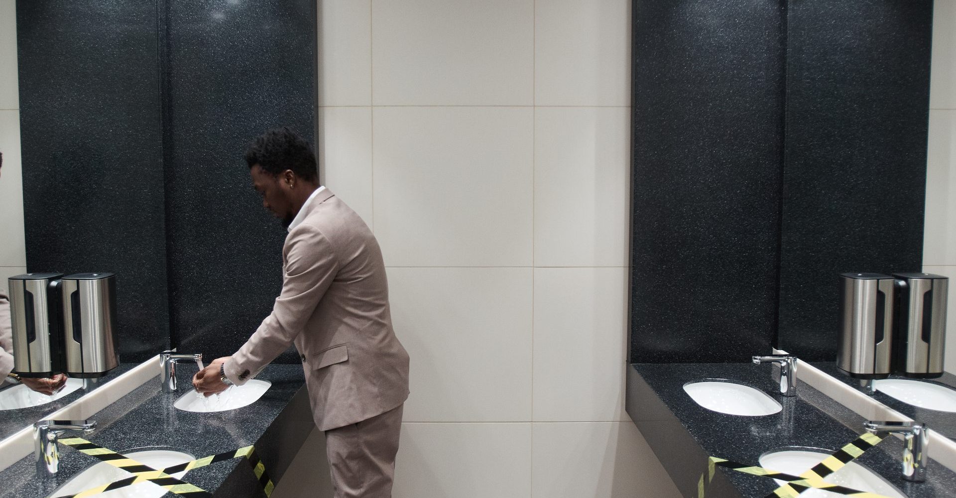 A man washes his hands in a corporate washroom with white sinks and dark walls.